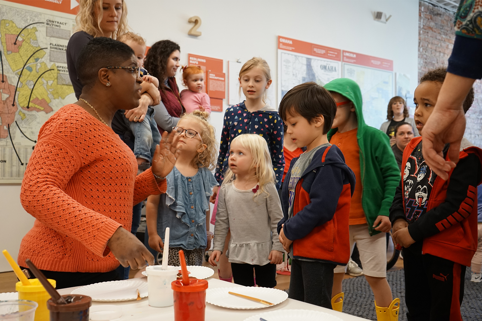 A group of toddlers listens closely to a teaching artist instructing them on finger painting