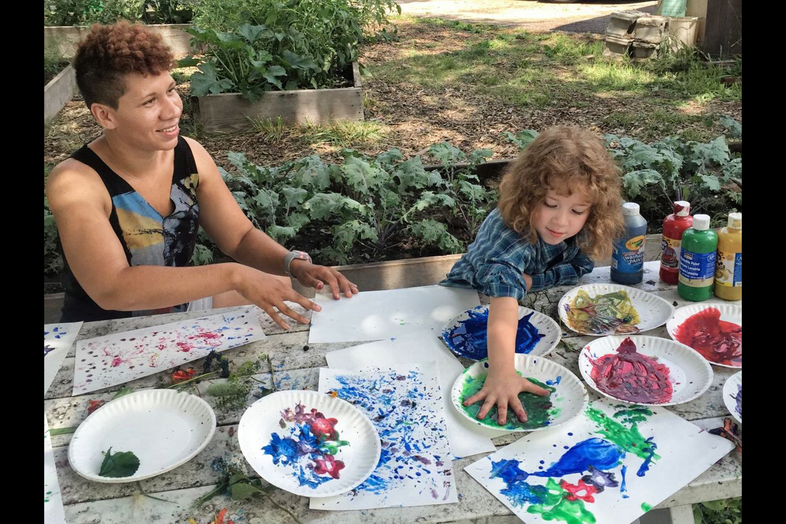 A teaching artist and young girl sit at a table outdoors full of finger paints