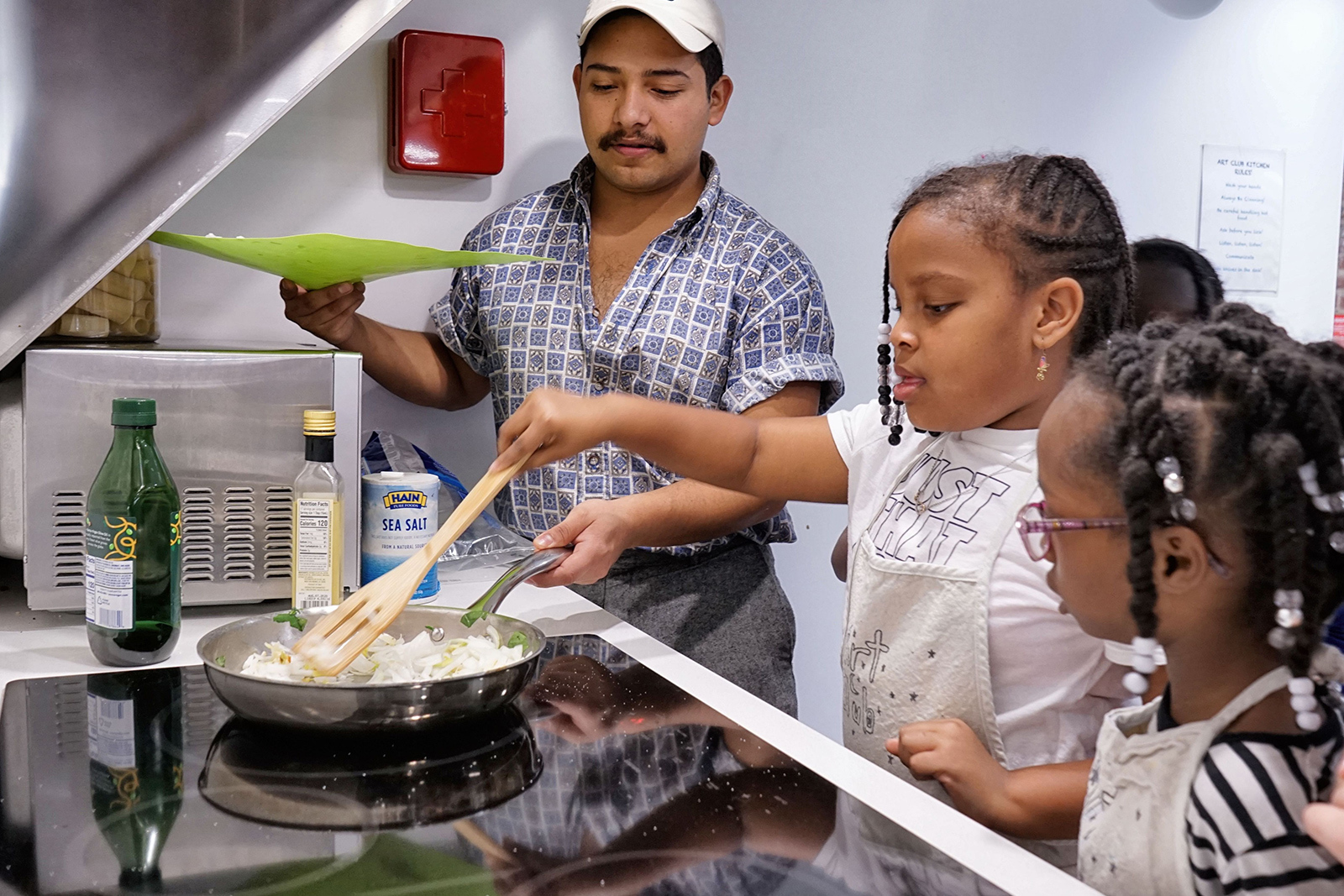 A teaching artist helps two young girls sautee onions in a pan