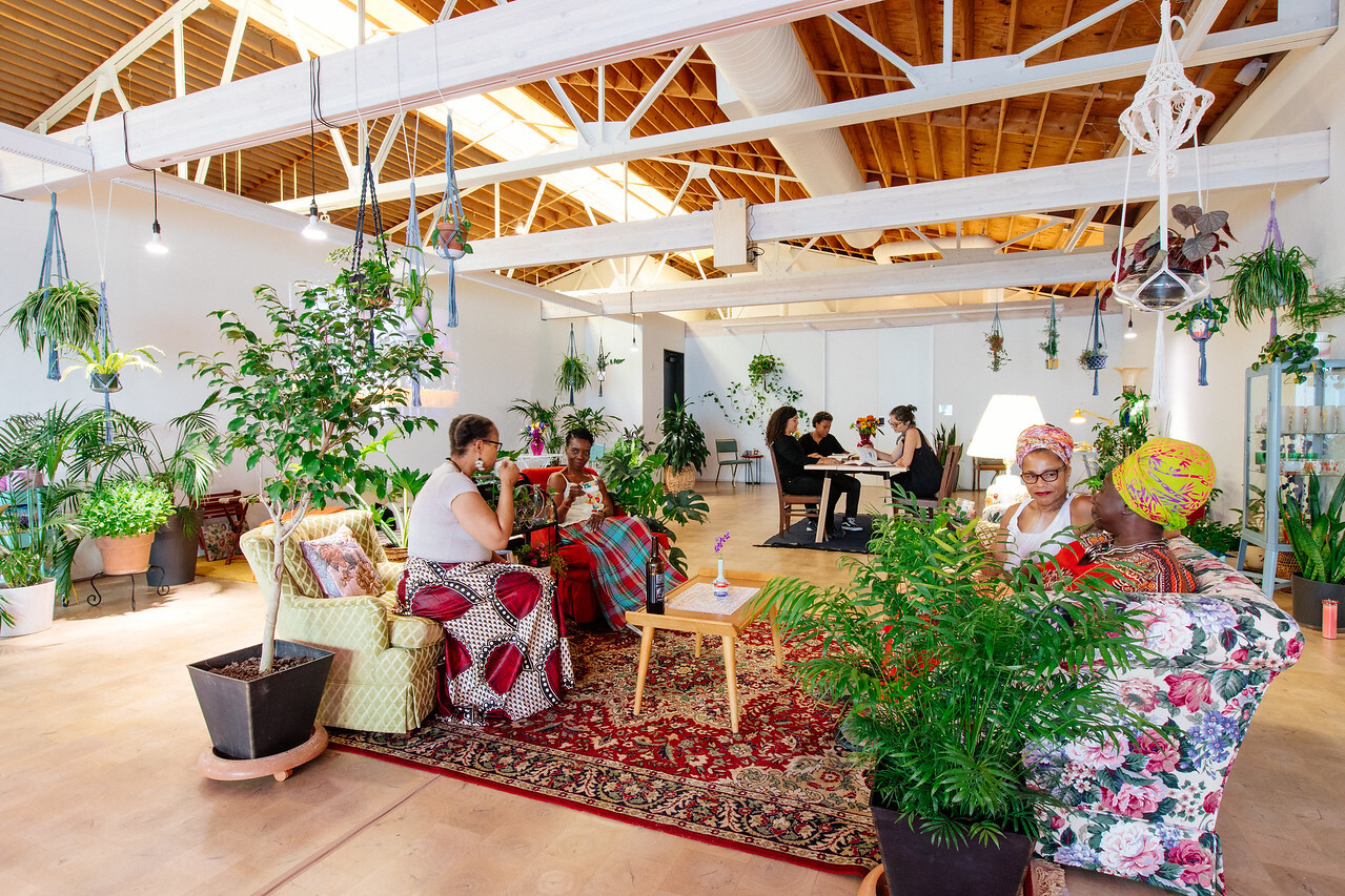 In the foreground, four Black women sit on floral and patterned couches around a small wooden table. The floor is carpeted, and the room is embellished with lush plants. In the background, three women sit in discussion at a wooden table.