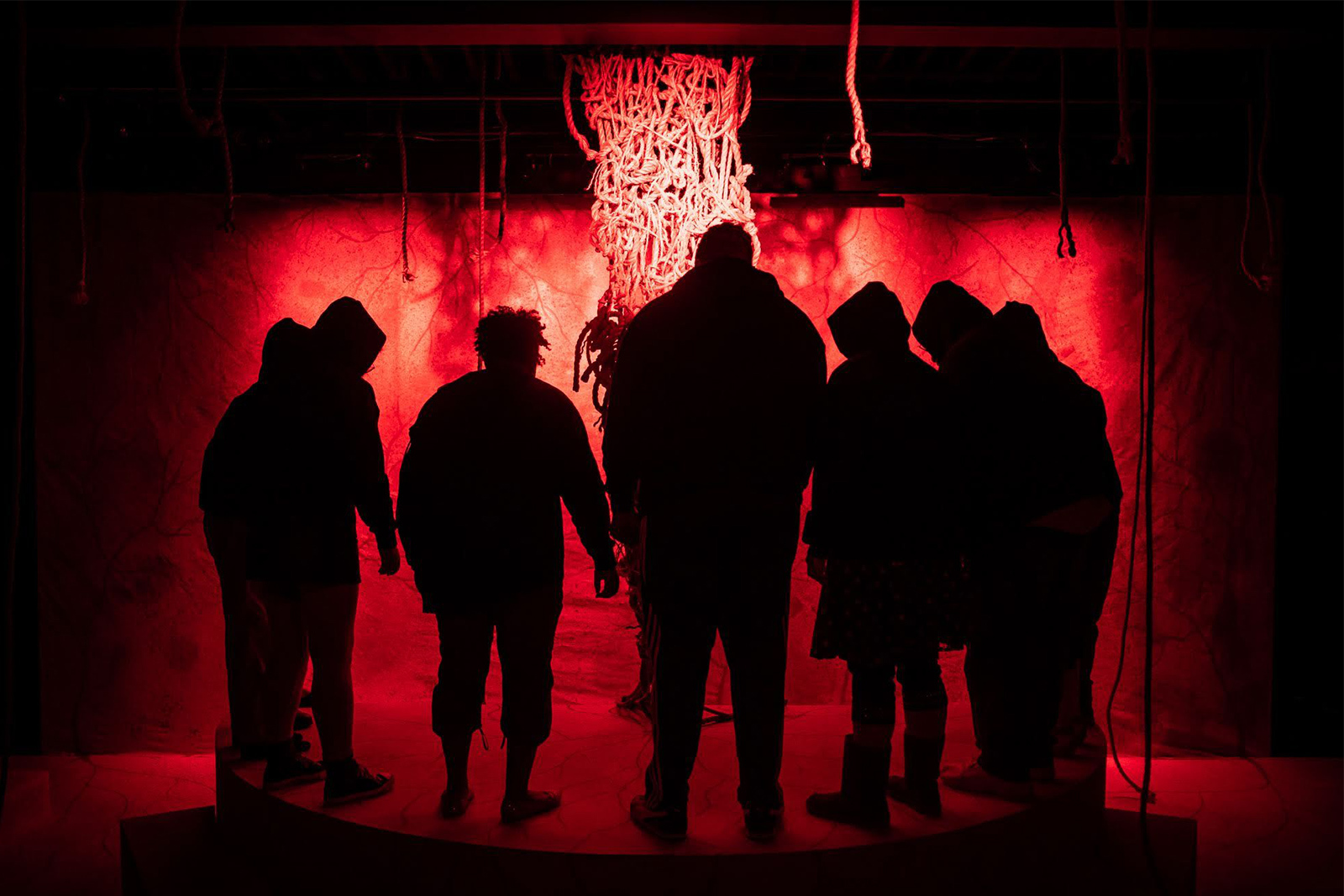 Blood at the Root cast of 7 stands center stage in low light heads bowed silhouetted against red light