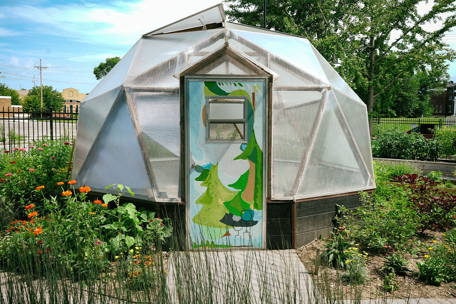 Abundance garden - photo of geodesic dome in Union garden. The garden is blooming with flowers and grasses and the door of the dome is painted with a bright, abstract nature scene.