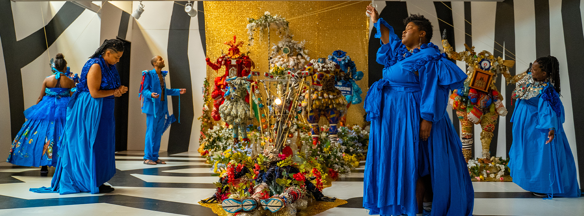Exhibitions banner - photo of artist Vanessa German in a blue dress with one arm raised standing in front a vibrant gold installation in her exhibition "sometimes.we.cannot.be.with.our.bodies."