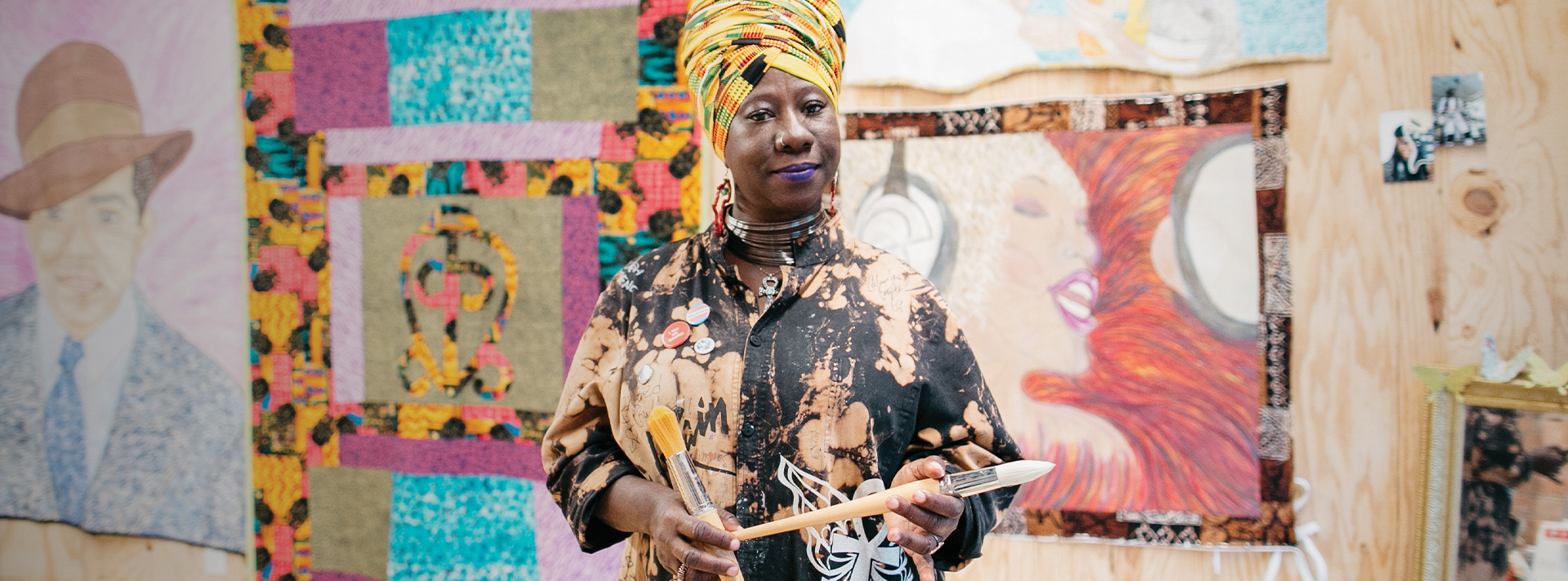 2017 Union Fellow Celeste Butler stands in front of two of her large quilts. She wears a yellow patterned head wrap and artist's smock, holding two paintbrushes. (Union fellowship header)